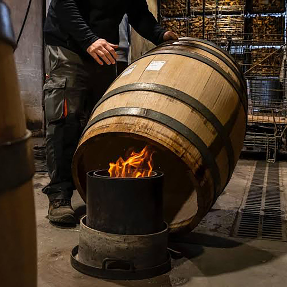 A person in dark clothing leans a large wooden barrel against a metal firebox with visible flames, in a rustic workshop or cellar setting with shelving and wood stacks in the background.