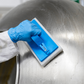 A close-up of a gloved hand using a blue and white scourer pad on a large stainless steel drum.