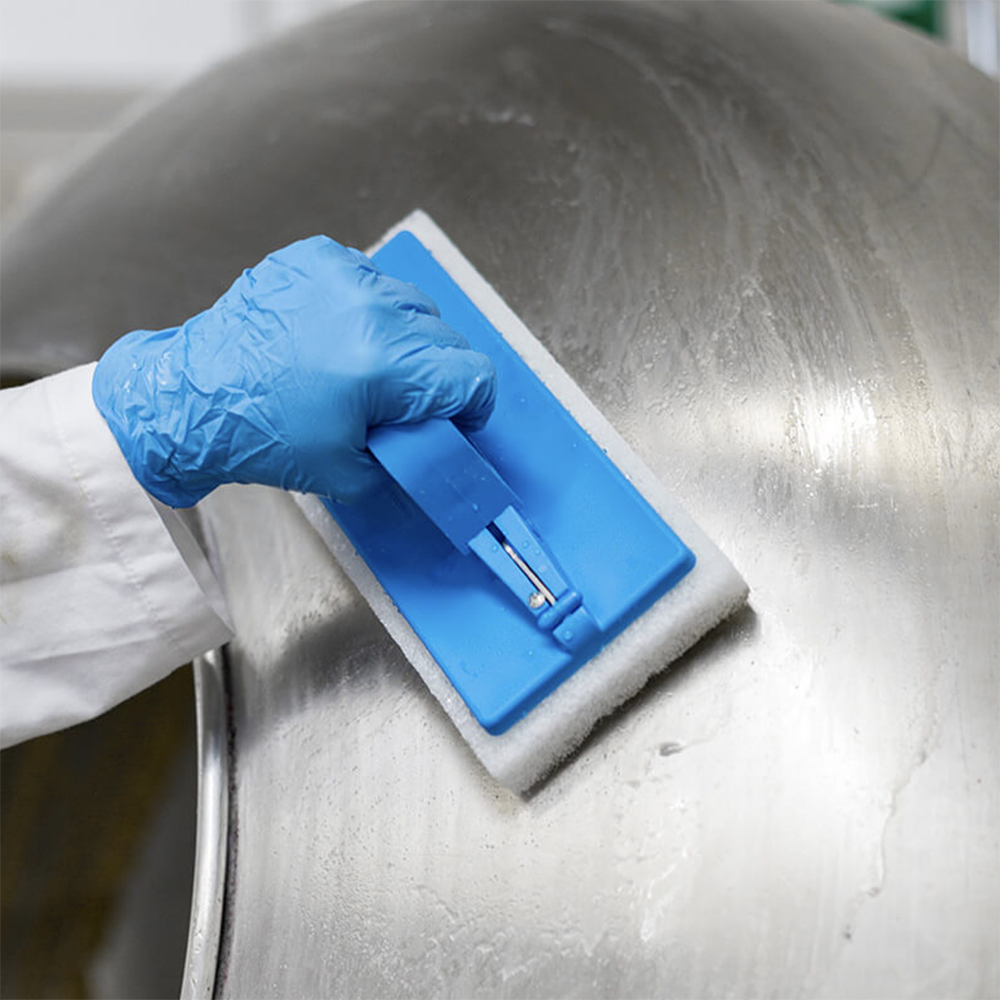 A close-up of a gloved hand using a blue and white scourer pad on a large stainless steel drum.